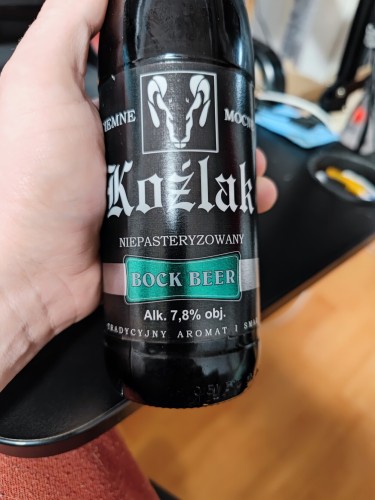 Butelka piwa trzymana w ręce, czarna etykieta "Koźlak", z zielonymi elementami i białymi napisami 