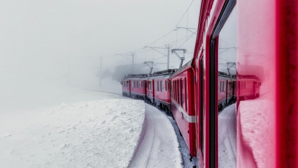 A view from the side window of a red electric train looking forward as the rails turn to the left. The front of the train begins to disappear into the whiteness of a blizzard. There is whiteness everywhere broken only by the redness of the train into the distance.


