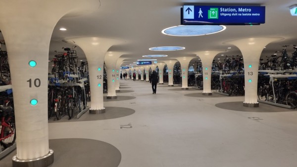Inside the underwater bike parking facility. On both sides of the central passage are pillars with row numbers on them. Behind the pillars, the rows with two-tiered bike racks begin. An information display hangs from the ceiling.