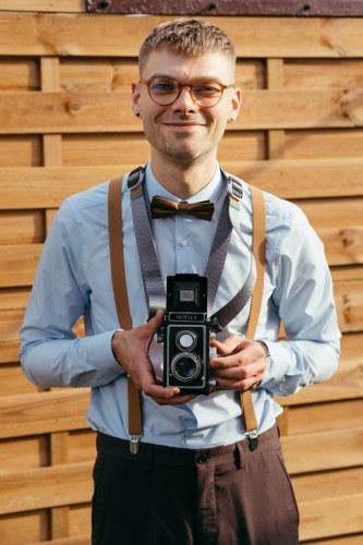 In the photo, I am wearing a long-sleeved shirt and fabric trousers. In addition, I am wearing glasses, a bow tie and braces. I am holding an old Zeiss Ikon Ikoflex analog camera hanging on a camera strap. In the background is a wooden fence.